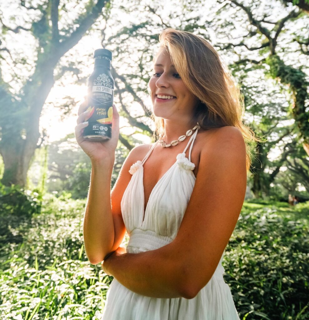 A woman wearing a white dress is holding a bottle of Karviva Ace Sport Mango.